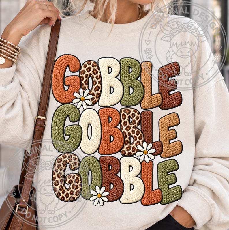 May include: A cream-colored crewneck sweatshirt featuring the text GOBBLE GOBBLE GOBBLE in a stacked design. The letters have a textured knit appearance in shades of orange, brown, green, and leopard print, accented with small white daisy flowers.