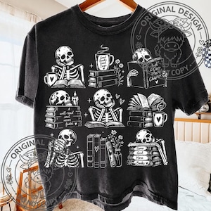 May include: A black short-sleeve t-shirt featuring a white graphic print of skeletons reading books and drinking from mugs. The design includes stacks of books, flowers, and text that reads Good Books Good Days, A well-read soul never fades, and Same Girl More Books.