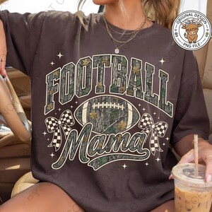 May include: A dark brown t-shirt featuring the text FOOTBALL MAMA in a camouflage pattern. The design includes a central football graphic, two checkered bows on the sides, and small star accents. A circular logo in the top right corner shows a highland cow.