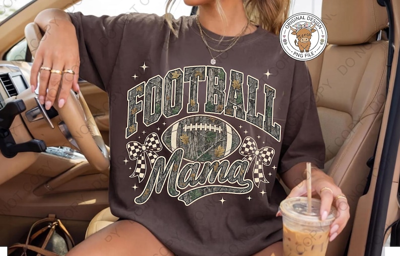 May include: A dark brown t-shirt featuring the text FOOTBALL MAMA in a camouflage pattern. The design includes a central football graphic, two checkered bows on the sides, and small star accents. A circular logo in the top right corner shows a highland cow.