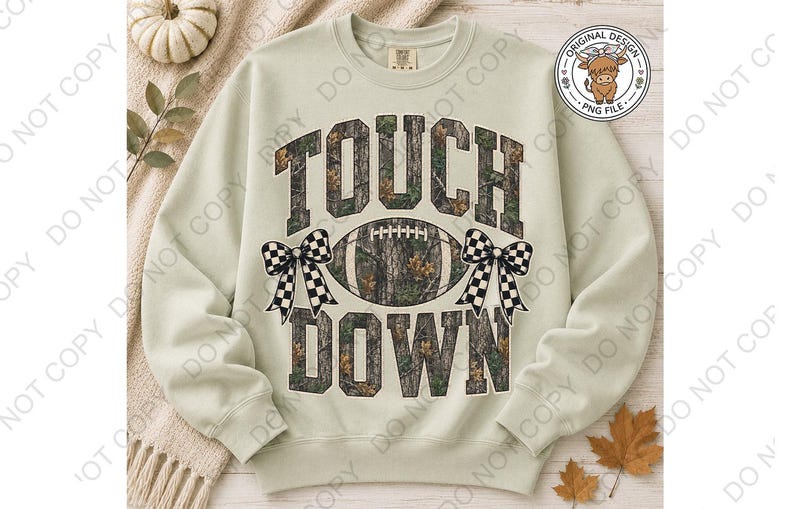 May include: A light sage green crewneck sweatshirt featuring the text TOUCH DOWN in a camouflage pattern. Between the words is a football graphic with a camouflage print, flanked by two black and white checkered bows on either side of the ball.