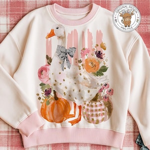 May include: A cream-colored sweatshirt with a pink trim features a whimsical design of a goose with a black bow, pumpkins, and floral accents. The design includes orange and pink pumpkins, and a floral arrangement. The sweatshirt is displayed on a plaid blanket.