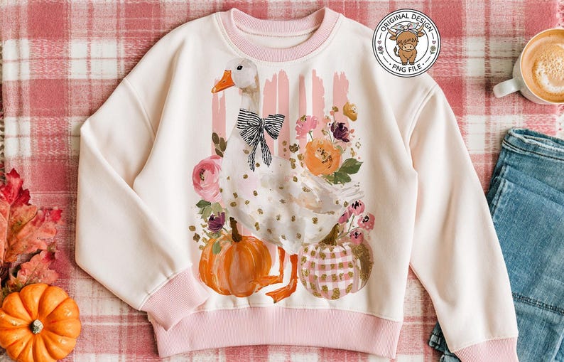 May include: A cream-colored sweatshirt with a pink trim features a whimsical design of a goose with a black bow, pumpkins, and floral accents. The design includes orange and pink pumpkins, and a floral arrangement. The sweatshirt is displayed on a plaid blanket.