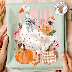 May include: A light green t-shirt featuring a white goose with a black bow tie, surrounded by pumpkins and flowers. The design includes the text "MOM" and a circular logo with a cow.