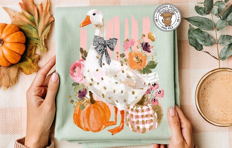 May include: A light green t-shirt featuring a white goose with a black bow tie, surrounded by pumpkins and flowers. The design includes the text "MOM" and a circular logo with a cow.