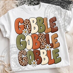 May include: A white short-sleeved t-shirt featuring the text GOBBLE GOBBLE GOBBLE printed in three rows. The letters have a textured knit appearance in shades of orange, brown, green, and leopard print, accented with small white daisy flowers.