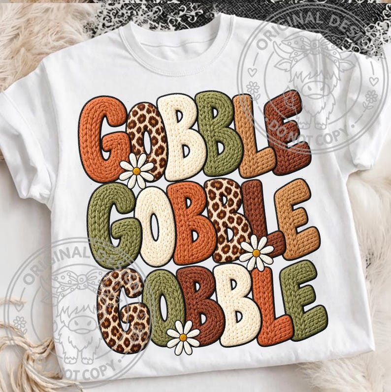 May include: A white short-sleeved t-shirt featuring the text GOBBLE GOBBLE GOBBLE printed in three rows. The letters have a textured knit appearance in shades of orange, brown, green, and leopard print, accented with small white daisy flowers.