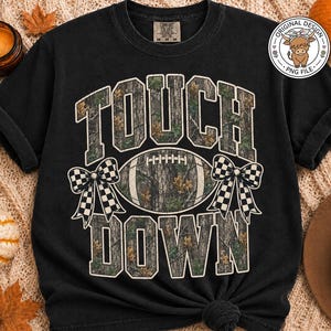 May include: A black short-sleeve t-shirt featuring the text TOUCH DOWN in a camouflage pattern. Between the words is a football graphic with a camouflage print, flanked by two black and white checkered bows. The shirt has a crew neckline.