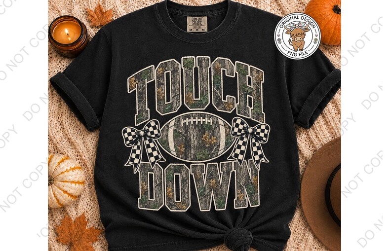 May include: A black short-sleeve t-shirt featuring the text TOUCH DOWN in a camouflage pattern. Between the words is a football graphic with a camouflage print, flanked by two black and white checkered bows. The shirt has a crew neckline.