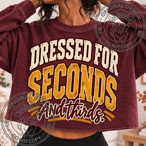 May include: A maroon long-sleeve cropped sweatshirt featuring the text DRESSED FOR SECONDS And thirds. The word DRESSED is in cream, SECONDS is in large yellow block letters, and And thirds is in a smaller white script font with yellow accents.
