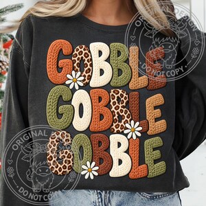 May include: A charcoal gray crewneck sweatshirt featuring the text GOBBLE GOBBLE GOBBLE in a chunky, knit-textured font. The letters alternate between solid orange, olive green, brown, cream, and leopard print patterns, accented by small white daisies.