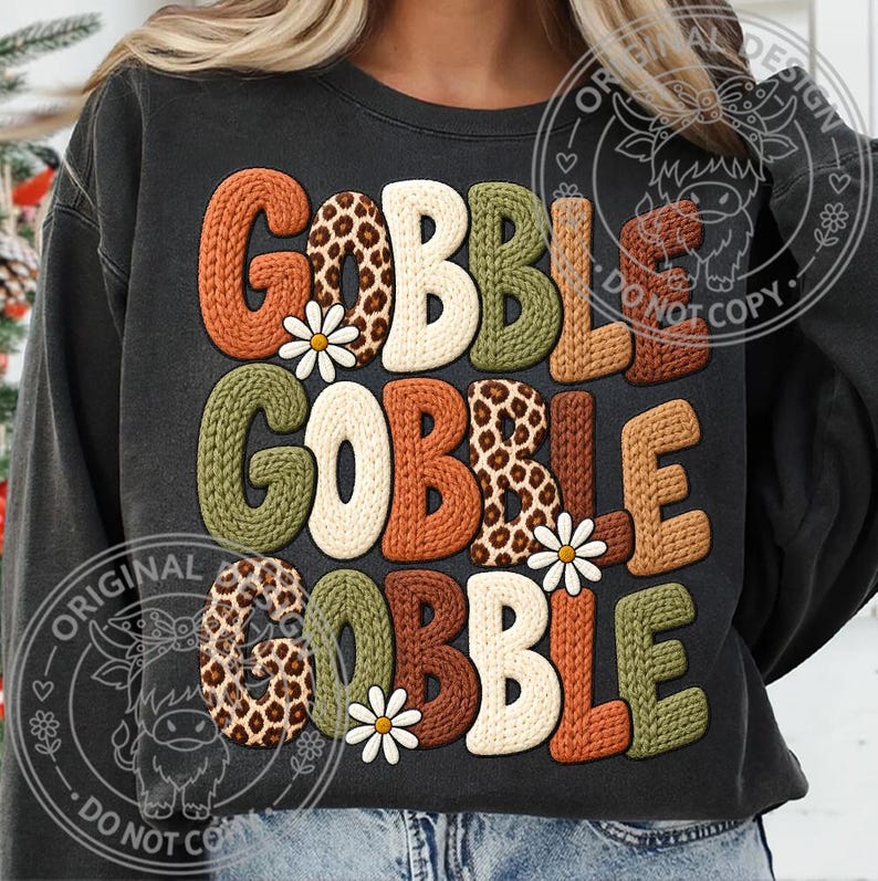 May include: A charcoal gray crewneck sweatshirt featuring the text GOBBLE GOBBLE GOBBLE in a chunky, knit-textured font. The letters alternate between solid orange, olive green, brown, cream, and leopard print patterns, accented by small white daisies.