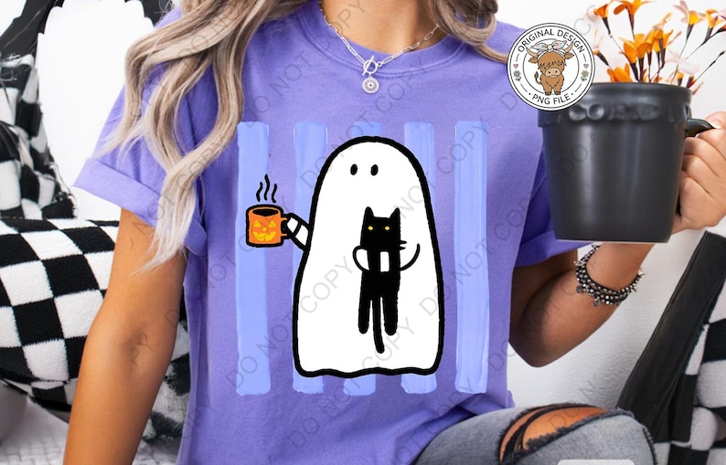 May include: A light purple t-shirt featuring a graphic of a white ghost holding an orange jack-o'-lantern mug with steam rising and a black cat. The design is set against vertical light blue stripes. The shirt has short sleeves and a crew neckline.
