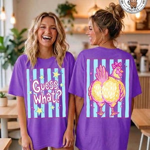 May include: Two women wearing purple t-shirts. The front of the shirt has the text "Guess What?" with stars and stripes. The back of the shirt features a cartoon chicken with a bow and necklace, set against a striped background.