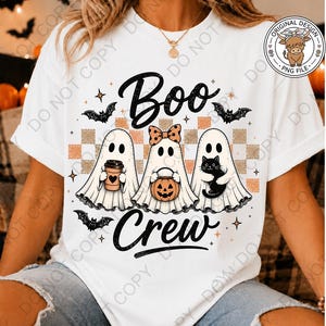May include: A white t-shirt featuring a graphic of three ghosts with the text "Boo Crew". The ghosts are positioned over a checkered pattern and are accompanied by bats, a coffee cup, a pumpkin bucket, and a black cat. The design includes stars.