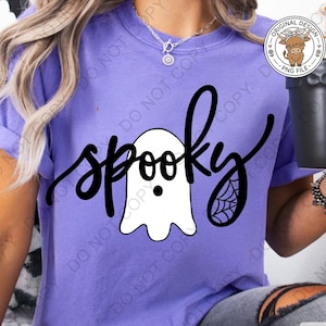 Op de afbeelding: Een paars t-shirt met korte mouwen en een zwarte afbeelding met het woord spooky in cursief schrift. Het ontwerp bevat een witte geestillustratie met zwarte ogen en mond, en een zwart spinnenwebdetail op de letter y.