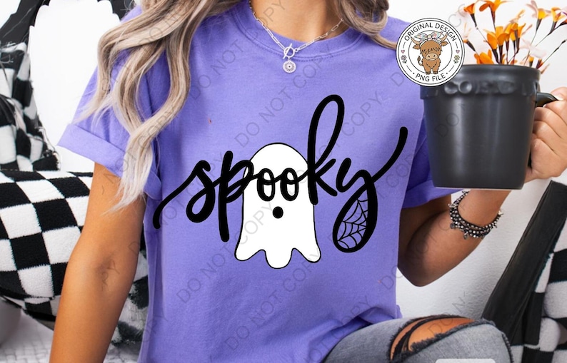 Op de afbeelding: Een paars t-shirt met korte mouwen en een zwarte afbeelding met het woord spooky in cursief schrift. Het ontwerp bevat een witte geestillustratie met zwarte ogen en mond, en een zwart spinnenwebdetail op de letter y.