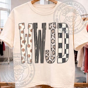 May include: A cream-colored short-sleeve t-shirt featuring the letters WWJD in a large, bold font. Each letter has a distinct pattern: the Ws feature brown polka dots and black vertical stripes, the J has a leopard print, and the D has a black and white checkerboard pattern.