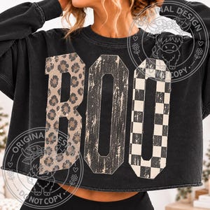May include: A black long-sleeve cropped sweatshirt featuring the word BOO printed in large block letters. The letter B has a brown leopard print, the letter O has a distressed black texture, and the second O has a black and white checkerboard pattern.