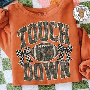 May include: A burnt orange long-sleeve sweatshirt featuring the text TOUCH DOWN in a camouflage pattern. A football with a camouflage texture is centered between the words, flanked by two black and white checkered bows on each side.