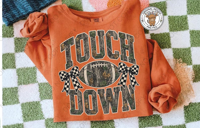 May include: A burnt orange long-sleeve sweatshirt featuring the text TOUCH DOWN in a camouflage pattern. A football with a camouflage texture is centered between the words, flanked by two black and white checkered bows on each side.