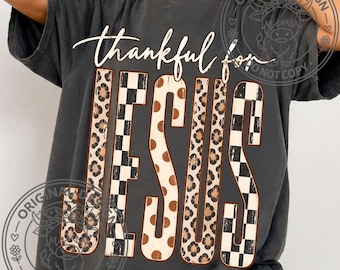 May include: A charcoal gray short-sleeve t-shirt featuring the white cursive text thankful for above the word JESUS in large block letters. The letters of JESUS are filled with alternating patterns of leopard print, black and white checkers, and brown polka dots.