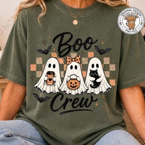 May include: An olive green t-shirt featuring a graphic of three white ghosts with the text "Boo Crew". The center ghost wears a polka dot bow, while the others hold a coffee cup, a jack-o'-lantern bucket, and a black cat. Bats and stars surround them.