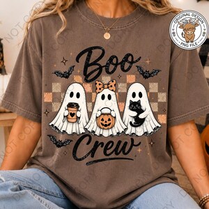 May include: A brown short-sleeve t-shirt featuring a graphic of three white ghosts with the text "Boo Crew". The ghosts are holding a coffee cup, a pumpkin bucket, and a black cat. The design includes bats and a checkered background.