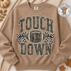 May include: A tan crewneck sweatshirt featuring the text TOUCH DOWN in a camouflage pattern. A football graphic with a camouflage print is centered between the words, flanked by two black and white checkered bows on either side of the ball.