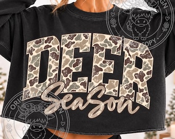 May include: A black cropped sweatshirt featuring the text "DEER" in large block letters filled with a tan, brown, and green camouflage pattern, with the word "SeaSon" written below in a tan cursive script font across the chest area.