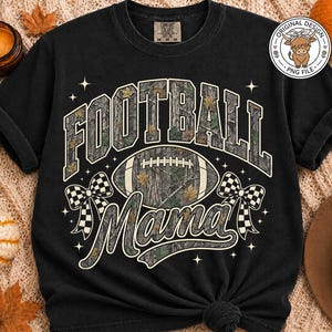 May include: A black short-sleeve t-shirt featuring a graphic print with the text "FOOTBALL Mama" in a camouflage pattern. The design includes a football, two checkered bows, and small star accents. The shirt has a crew neckline and a relaxed fit.