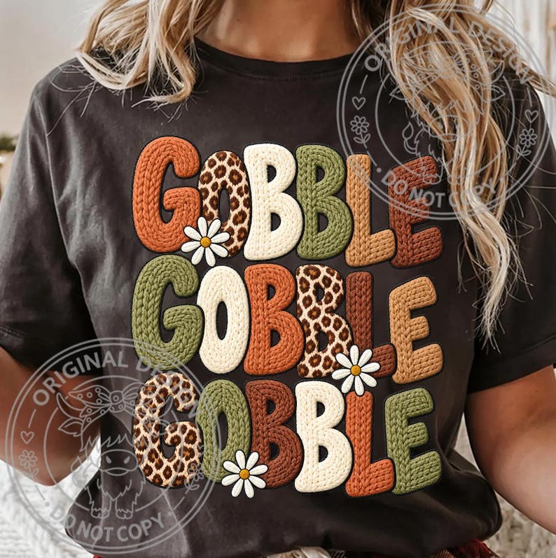 May include: A dark gray t-shirt featuring the text GOBBLE GOBBLE GOBBLE in a stacked design. The letters have a textured knit appearance in orange, cream, olive green, brown, and leopard print, accented with small white daisy flower graphics.