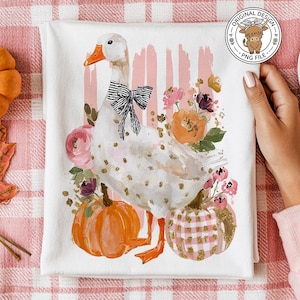 May include: White tea towel featuring a watercolor goose with a black bow, surrounded by pumpkins and flowers. The design includes pink and gold accents, perfect for fall decor.