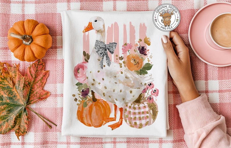May include: White tea towel featuring a watercolor goose with a black bow, surrounded by pumpkins and flowers. The design includes pink and gold accents, perfect for fall decor.