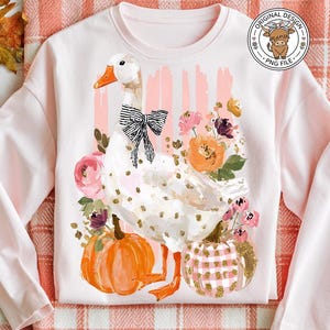 May include: A light pink sweatshirt featuring a whimsical goose design with a black bow, surrounded by flowers and pumpkins. The design includes gold accents and is set against a backdrop of pink stripes. The image also includes a pumpkin, fall leaves, and a pair of blue jeans.