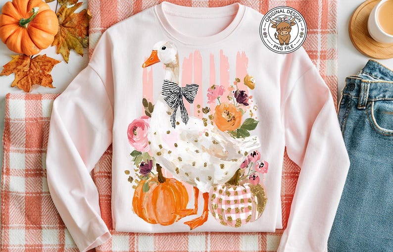 May include: A light pink sweatshirt featuring a whimsical goose design with a black bow, surrounded by flowers and pumpkins. The design includes gold accents and is set against a backdrop of pink stripes. The image also includes a pumpkin, fall leaves, and a pair of blue jeans.