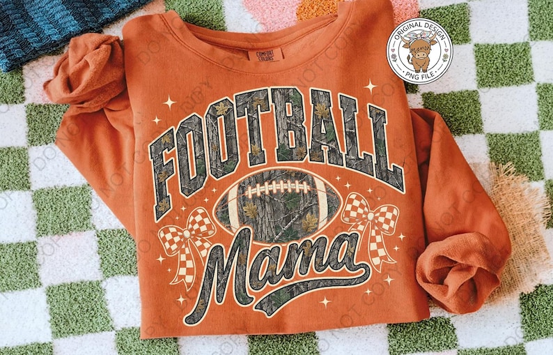 May include: An orange long-sleeve sweatshirt featuring a graphic print with the text "FOOTBALL MAMA" in a camouflage pattern. The design includes a football, two checkered bows, and small star accents. A circular logo is visible in the top right corner.