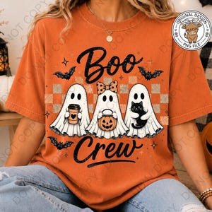 May include: An orange short-sleeve t-shirt featuring a graphic of three white ghosts with the text "Boo Crew". The ghosts are holding a coffee cup, a pumpkin bucket, and a black cat. The design includes small black bats and a checkered background.