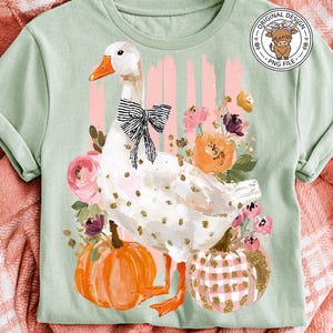 May include: A light green t-shirt featuring a whimsical goose design with a black bow, gold polka dots, and orange feet. The goose is surrounded by flowers and pumpkins. The shirt has a round "Original Design" logo.