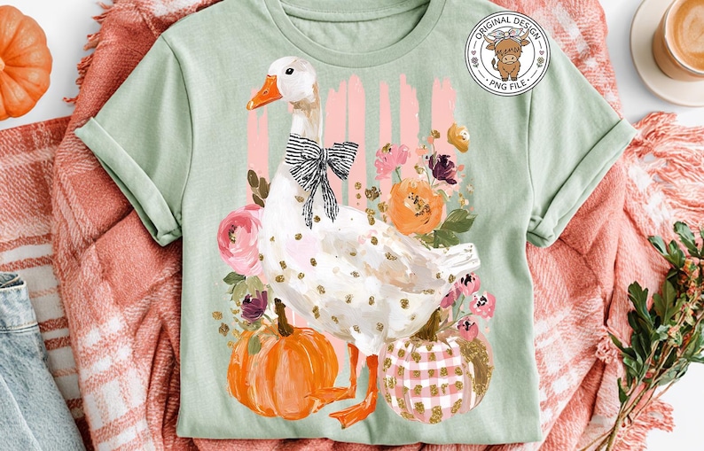 May include: A light green t-shirt featuring a whimsical goose design with a black bow, gold polka dots, and orange feet. The goose is surrounded by flowers and pumpkins. The shirt has a round "Original Design" logo.