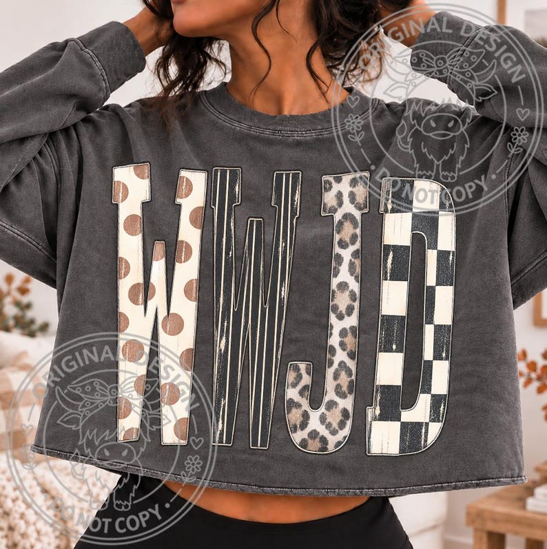 May include: A charcoal gray long-sleeve cropped sweatshirt featuring the letters WWJD in large, decorative block fonts. Each letter displays a unique pattern: polka dots, vertical stripes, leopard print, and a black and white checkerboard.