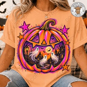 May include: A peach-colored t-shirt featuring a large, vibrant graphic of a jack-o'-lantern. Inside the pumpkin, a spooky scene depicts three white ghosts, a full moon, bats, and a haunted house. The design is accented with bright pink stars.
