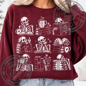 May include: A maroon crewneck sweatshirt featuring a white graphic print of nine distinct scenes with skeletons, stacks of books, mugs, and flowers. The text includes phrases like Good Books Good Days, A well-read soul never fades, and Same Girl More Books.