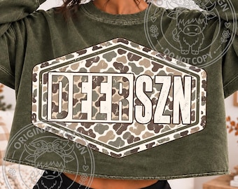 May include: An olive green long-sleeve sweatshirt featuring a large, central hexagonal graphic. The graphic contains a camouflage pattern in shades of brown, tan, and green, with the white text DEERSZN printed across the center in a bold font.