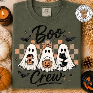 May include: An olive green crewneck t-shirt featuring a graphic of three ghosts with the text "Boo Crew". The ghosts are holding a coffee cup, a pumpkin bucket, and a black cat. The design includes a checkered background, bats, and stars.