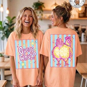 May include: Peach-colored t-shirts with a graphic design. The front of the shirt says "Guess What?" in a playful font, surrounded by stars. The back features a cartoon chicken with a pink bow and necklace.