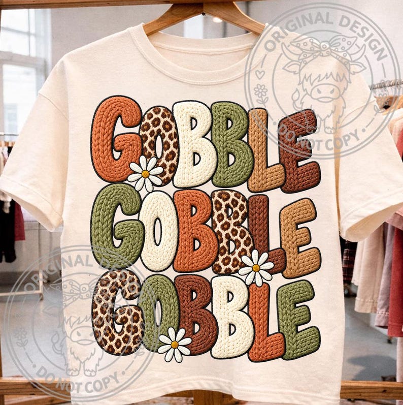 May include: A cream-colored t-shirt featuring the text GOBBLE GOBBLE GOBBLE in a chunky, knit-textured font. The letters alternate between solid orange, olive green, brown, and leopard print patterns, accented by three small white daisy flowers.