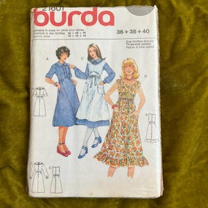 May include: A vintage Burda sewing pattern packet featuring illustrations of three dresses. The packet has the Burda logo in red and white, with text in English and German. The dresses are in blue, white, and floral patterns.