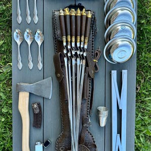 Puede incluir: Un conjunto de utensilios de cocina para exteriores, que incluye brochetas, un cuchillo, un hacha y pequeños cuencos de metal. Las brochetas tienen mangos decorativos y se guardan en una funda de cuero. También se incluyen tenedores, cucharas y una pequeña taza de metal.