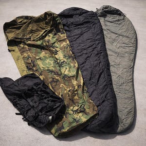 May include: Four sleeping bags are shown, including a camouflage print, black, and olive green. A black stuff sack is also visible. The items are arranged on a grey surface. The sleeping bags are in various states of being open.